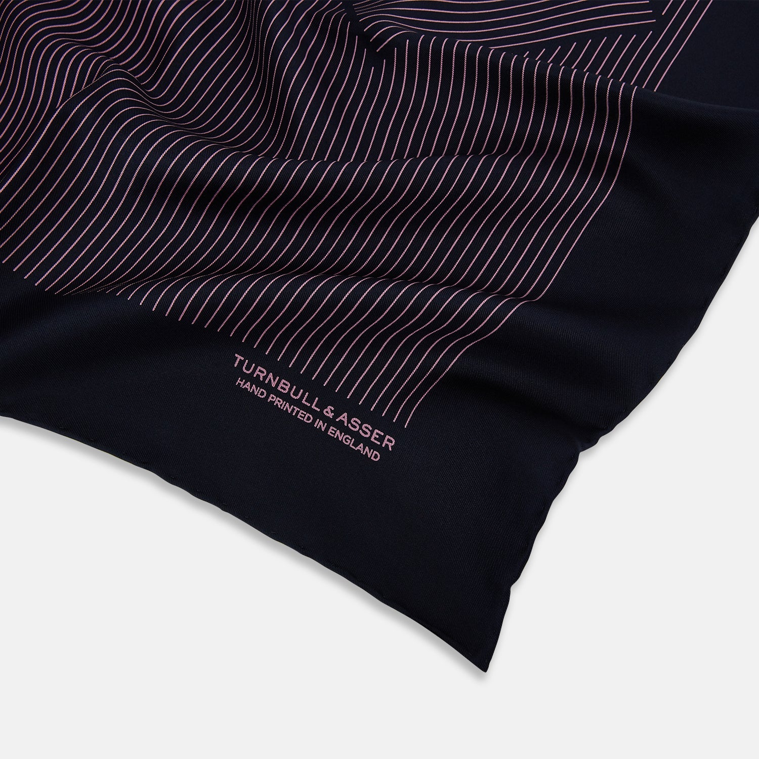 View 2 of product The Navy and Pink Stripe Silk Pocket Square by Turnbull & Asser features thin pink stripes, a geometric border, hand-rolled edges, and "TURNBULL & ASSER HAND PRINTED IN ENGLAND" printed in white.