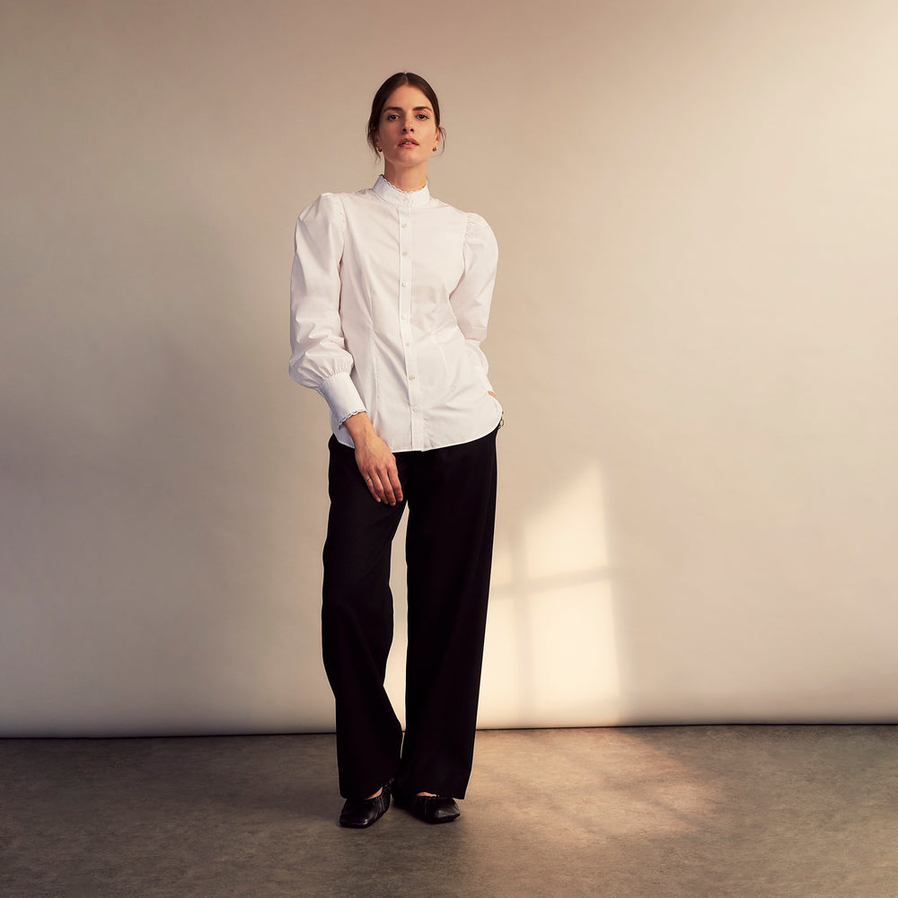 A woman stands in profile, looking down with hands in her pockets, wearing the Turnbull & Asser Turnbull and The Deck White Cotton Expressionist Shirt, paired with high-waisted black-and-white houndstooth pants.
