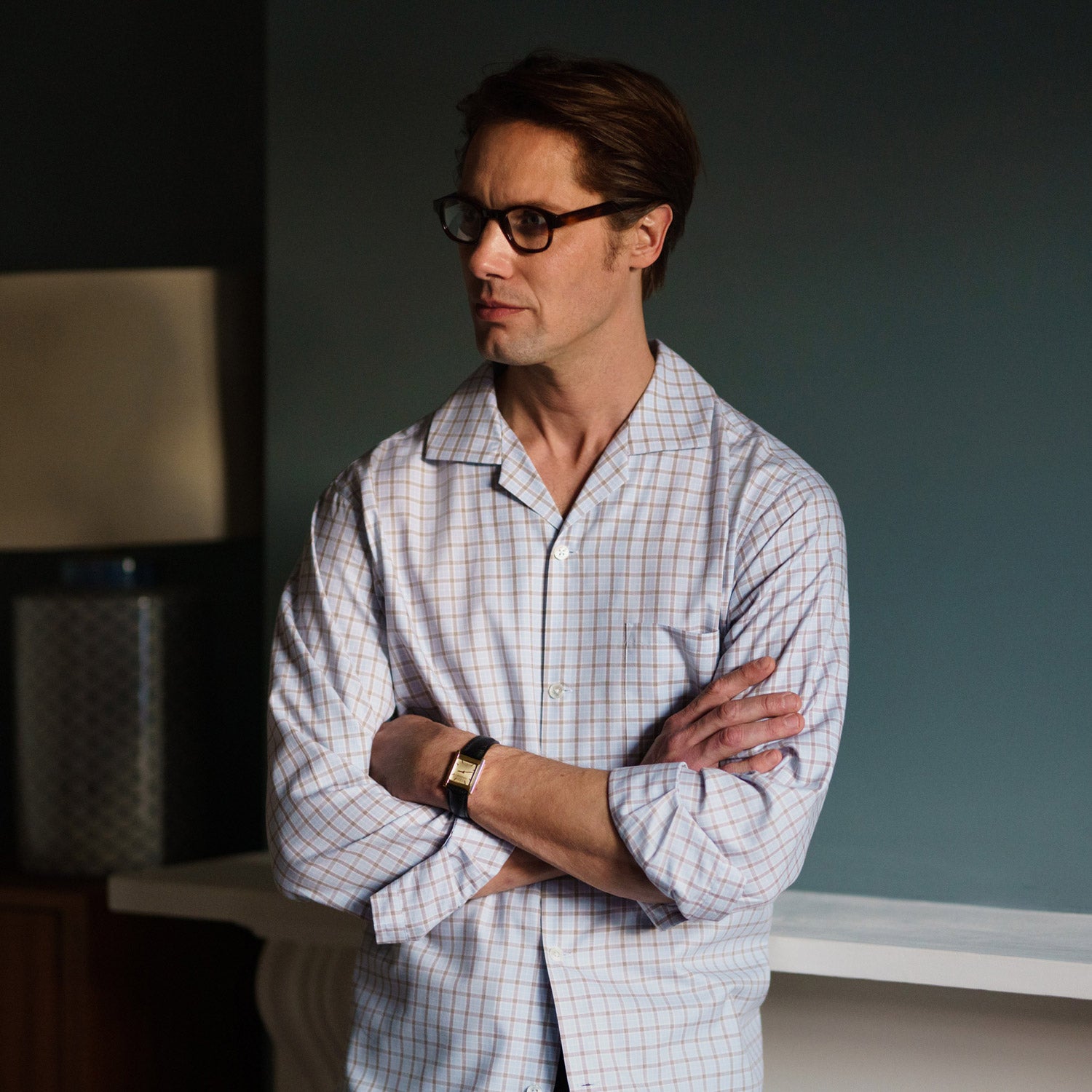 View 5 of product A man wearing glasses and a Pale Blue Multi Check Cashmerello Holiday Fit Winnington Shirt by Turnbull & Asser leans casually against a white countertop, looking at the camera with a neutral expression.