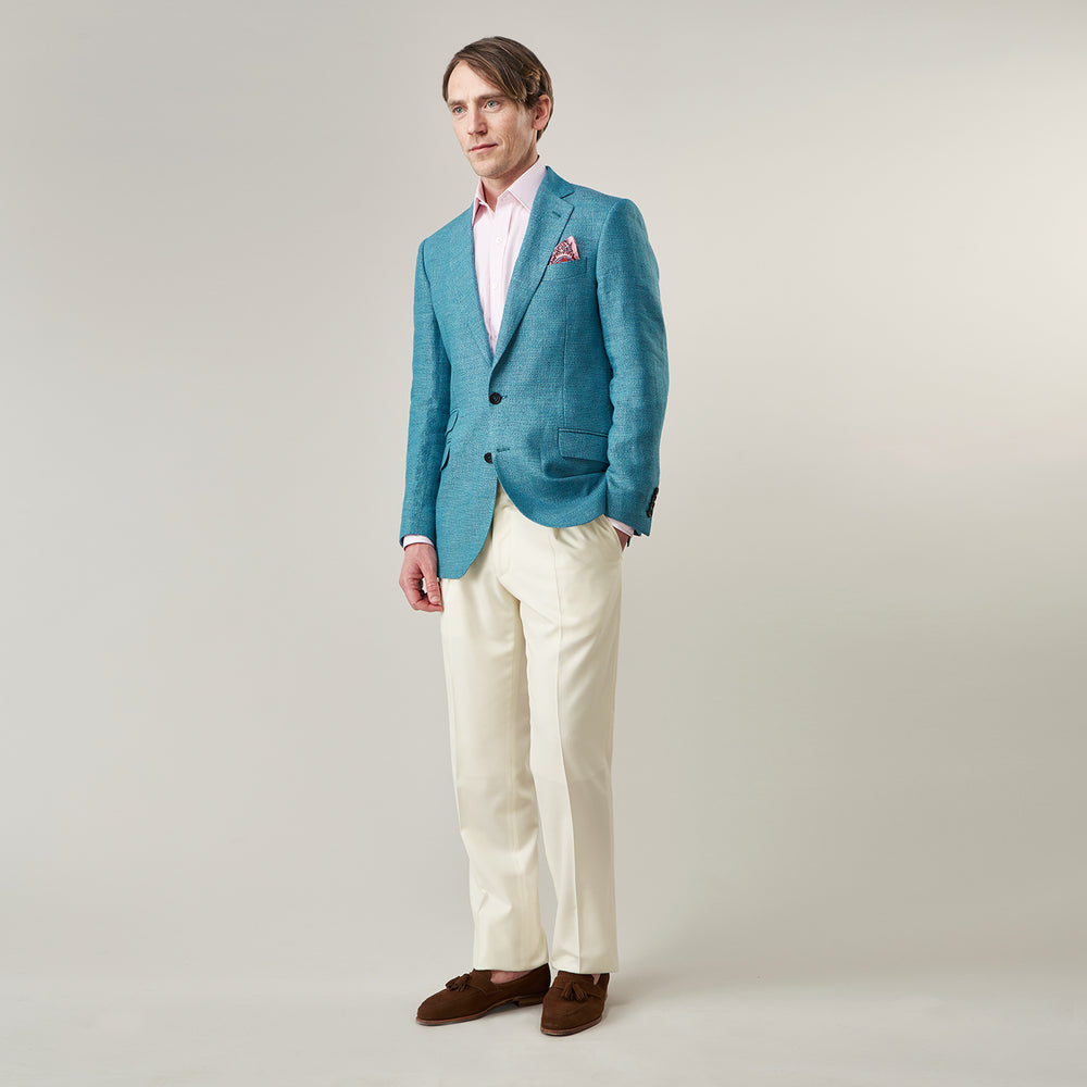A man with short brown hair and a neutral expression stands against a plain background, wearing a Teal Linen Barrington Blazer by Turnbull & Asser, a light pink dress shirt, white trousers, brown loafers, and a patterned pocket square.