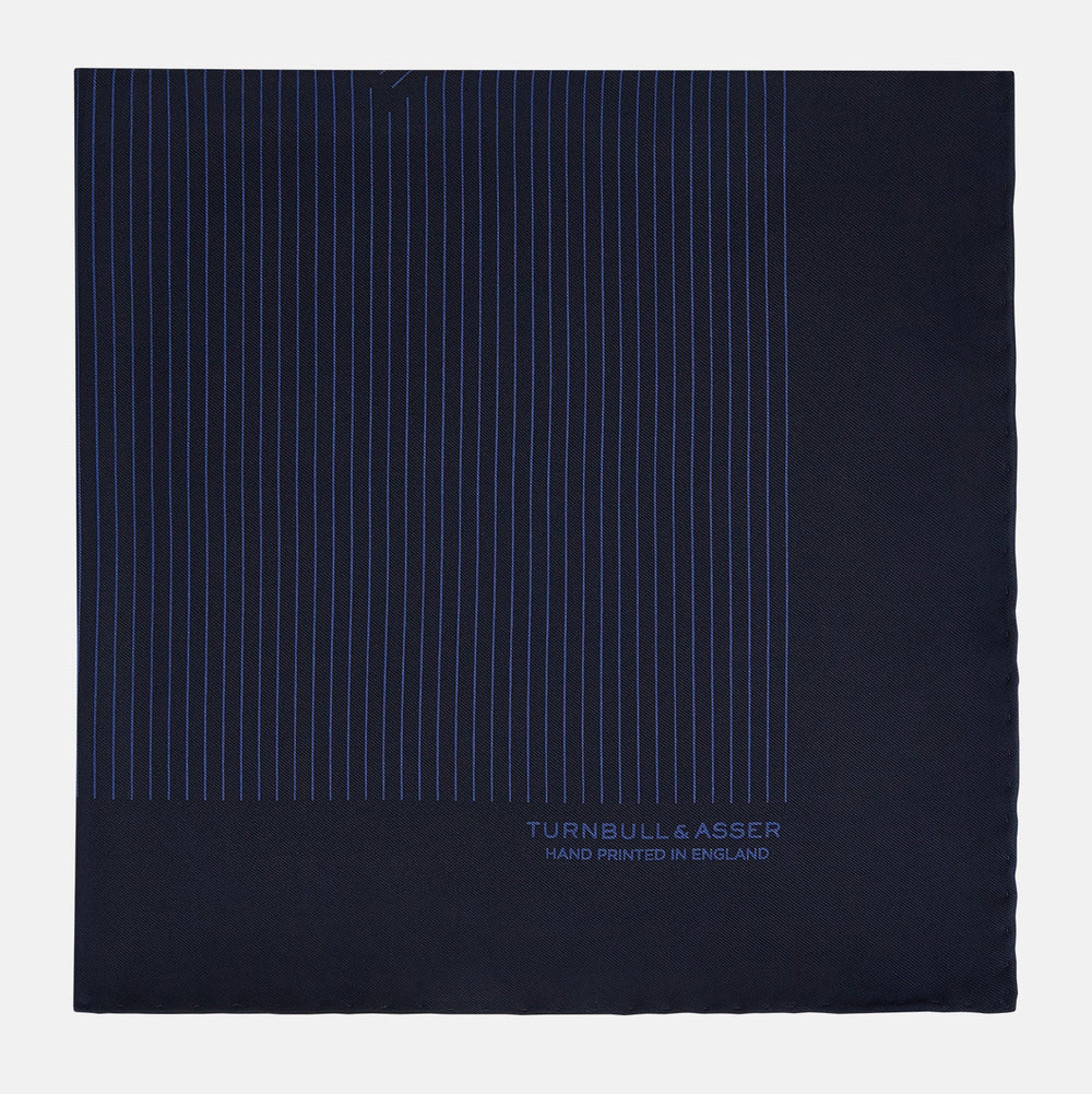 The Navy Stripe Silk Pocket Square by Turnbull & Asser features a dark navy silk with thin light blue stripes and hand-rolled edges, folded to reveal both sides and “TURNBULL & ASSER HAND PRINTED IN ENGLAND” in the corner.