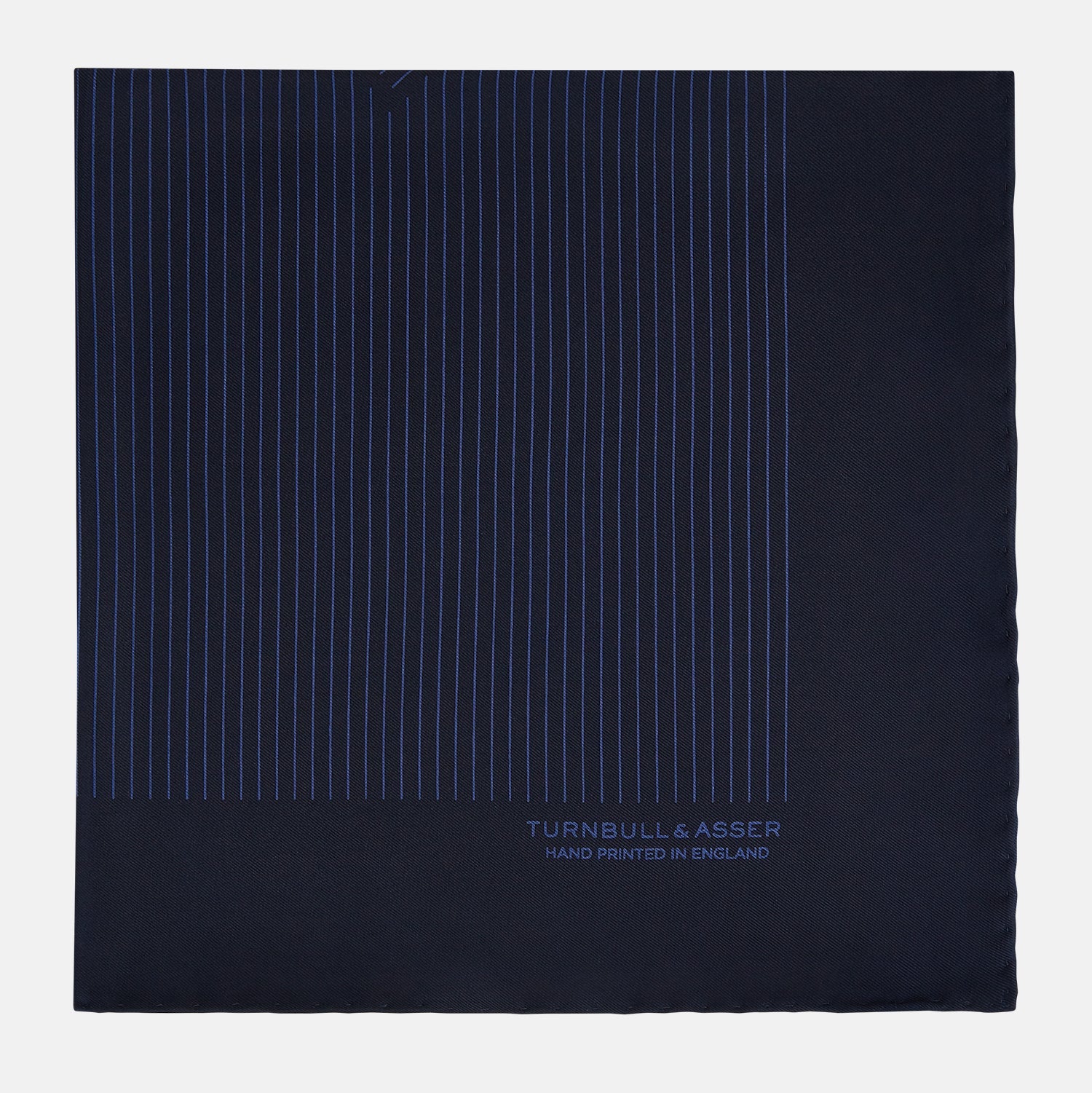 View 4 of product The Navy Stripe Silk Pocket Square by Turnbull & Asser features a dark navy silk with thin light blue stripes and hand-rolled edges, folded to reveal both sides and “TURNBULL & ASSER HAND PRINTED IN ENGLAND” in the corner.