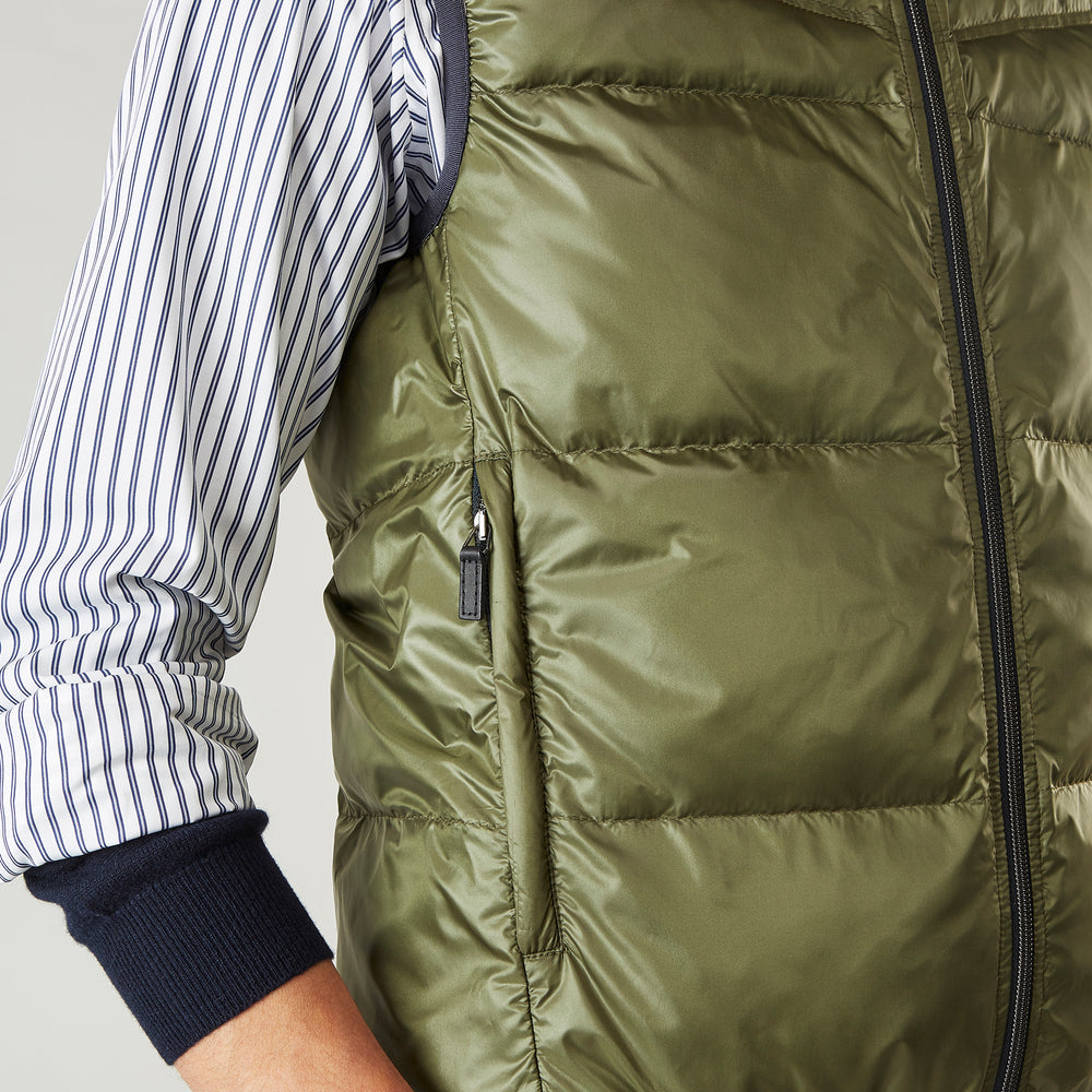 A man stands against a plain background wearing the Turnbull & Asser Green Peyton Sealup Quilted Gilet over a striped shirt, dark trousers, and white sneakers, with his hands in his pockets and looking slightly to the side.