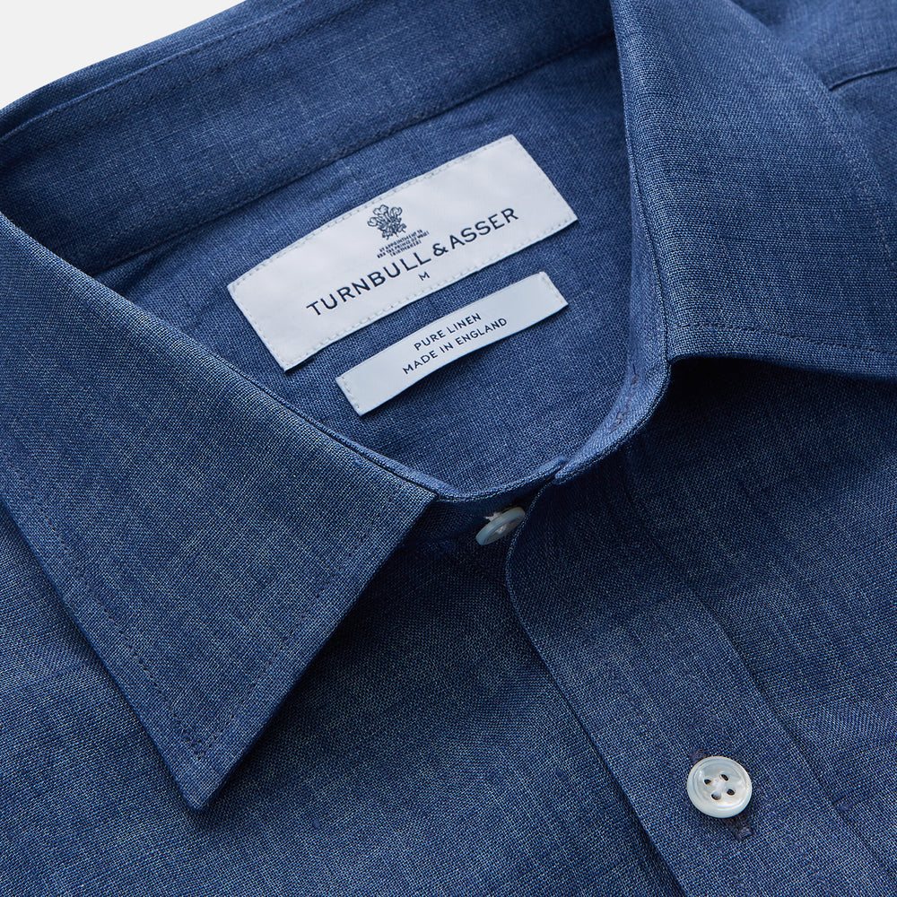 The Blue Weekend Fit Linen Finch Shirt by Turnbull & Asser features a Derby collar, chest pocket, and white buttons. Made from mid-blue linen chambray, it offers effortless casual style when laid flat on a plain white background.