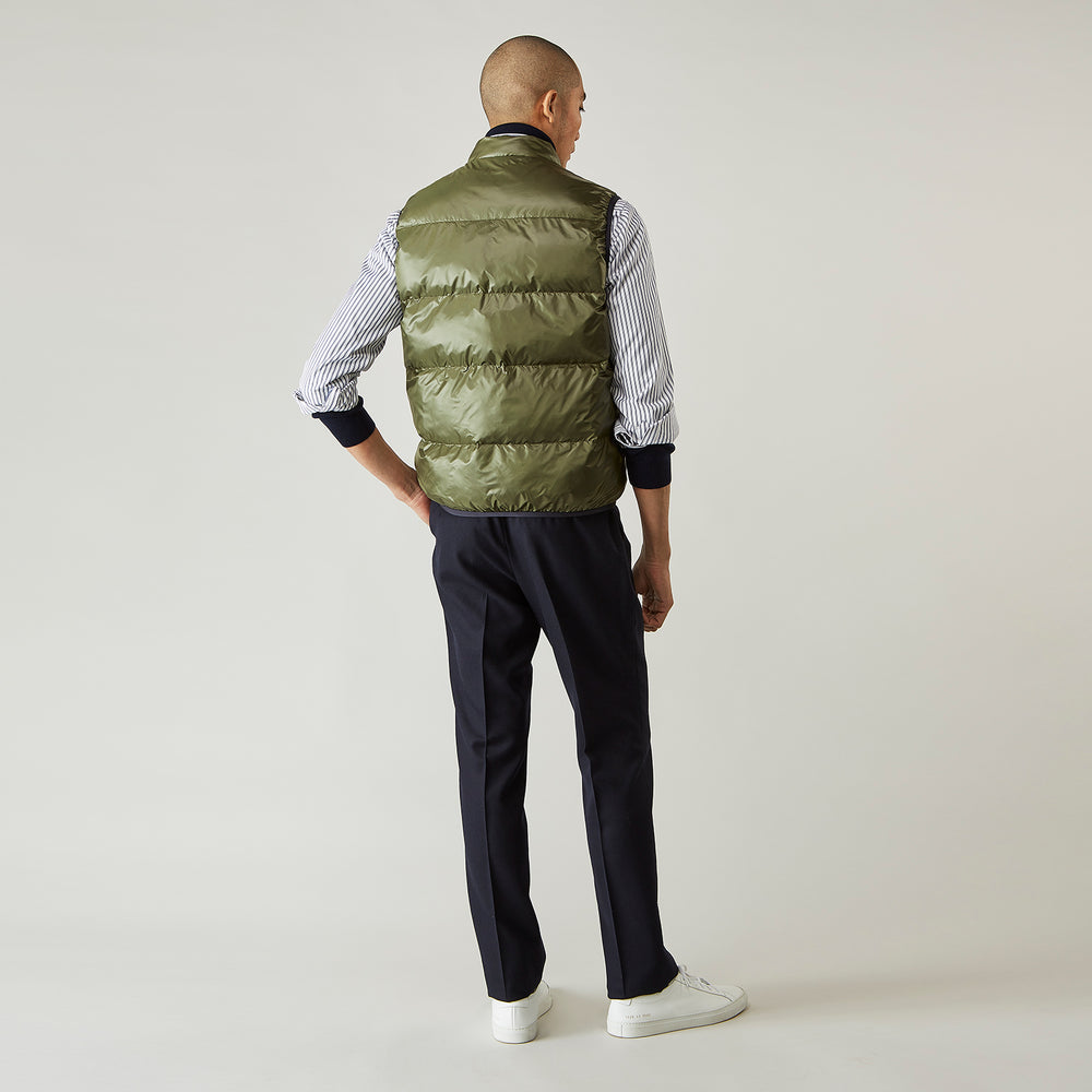A man stands against a plain background wearing the Turnbull & Asser Green Peyton Sealup Quilted Gilet over a striped shirt, dark trousers, and white sneakers, with his hands in his pockets and looking slightly to the side.
