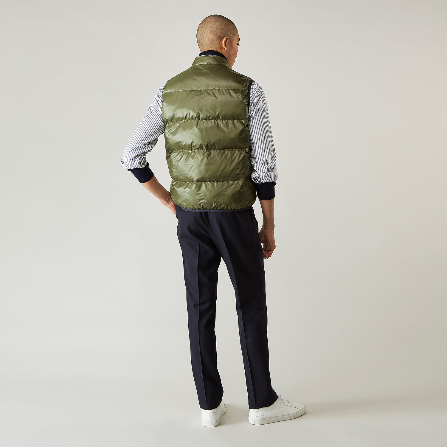 View 4 of product A man stands against a plain background wearing the Turnbull & Asser Green Peyton Sealup Quilted Gilet over a striped shirt, dark trousers, and white sneakers, with his hands in his pockets and looking slightly to the side.