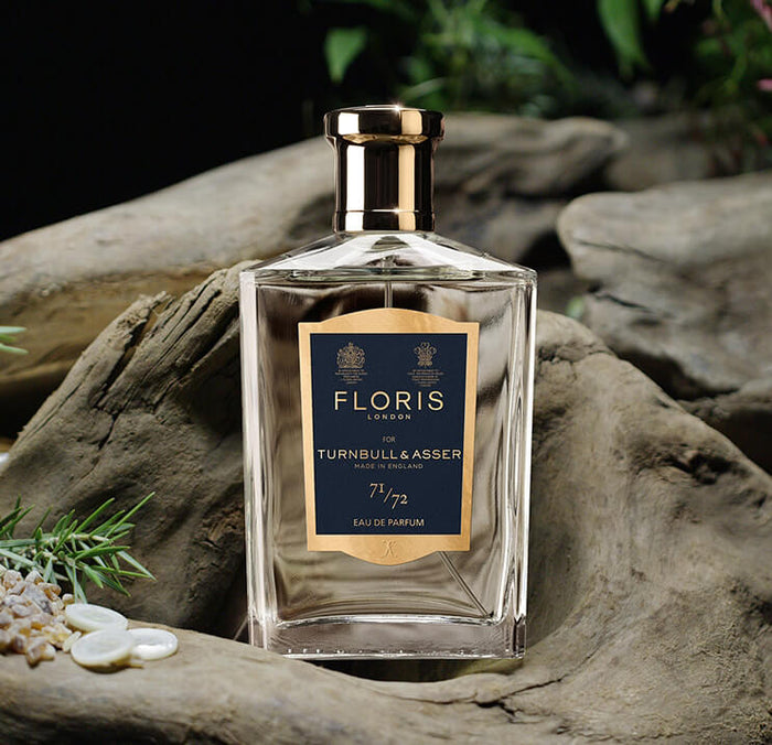 A bottle of Floris London Turnbull & Asser Eau de Parfum sits on a piece of driftwood, surrounded by greenery and small white stones, with a gold and navy label on the front.