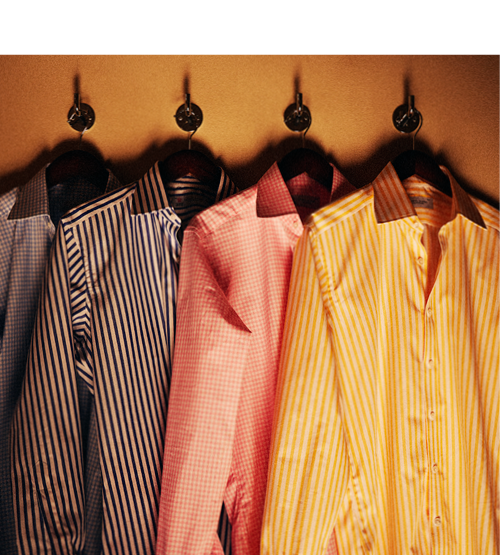Four dress shirts in blue stripes, black stripes, pink checks, and yellow stripes hang on wooden hangers from hooks against a warm-colored wall.