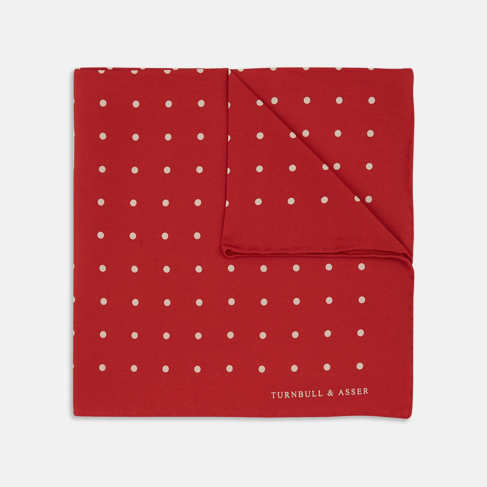The Red and White Spot Silk Pocket Square by Turnbull & Asser features evenly spaced white polka dots on red silk with the brand's name printed in one corner. A folded edge reveals the reverse of this timeless pocket square.