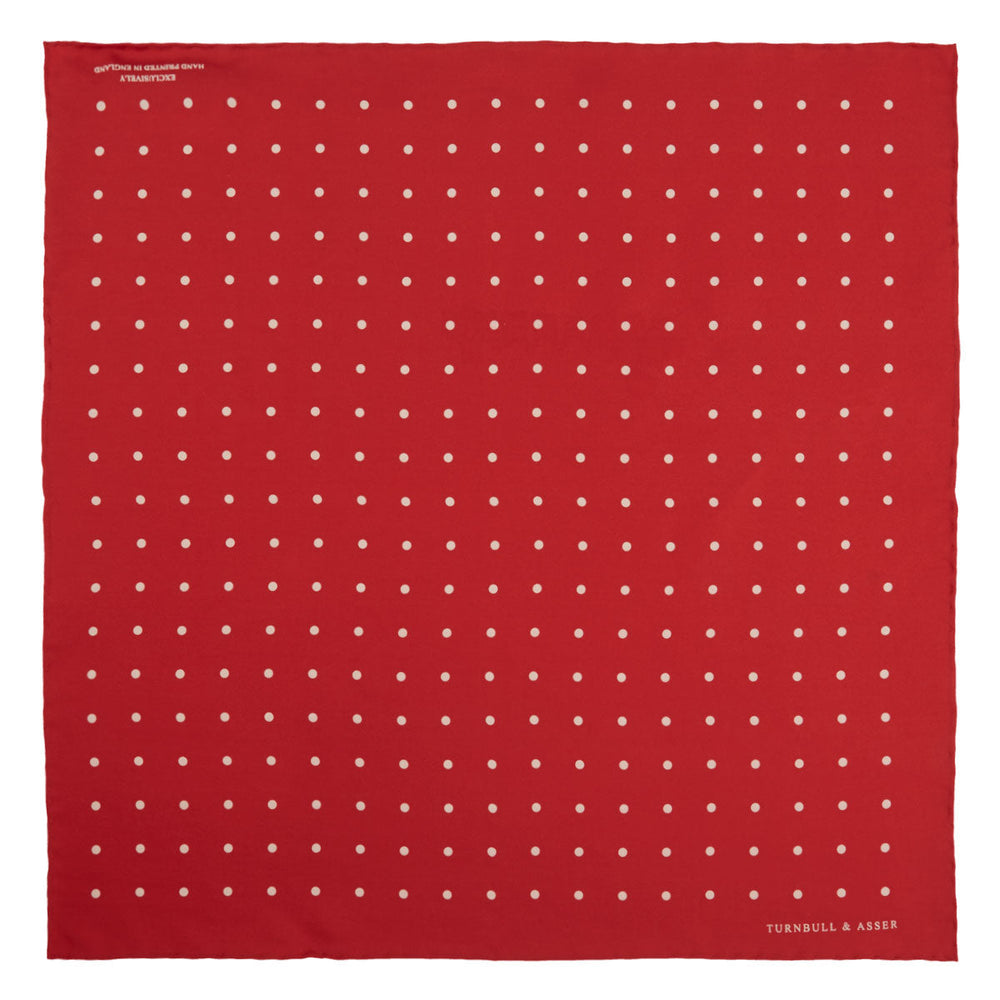 The Red and White Spot Silk Pocket Square by Turnbull & Asser features evenly spaced white polka dots on red silk with the brand's name printed in one corner. A folded edge reveals the reverse of this timeless pocket square.
