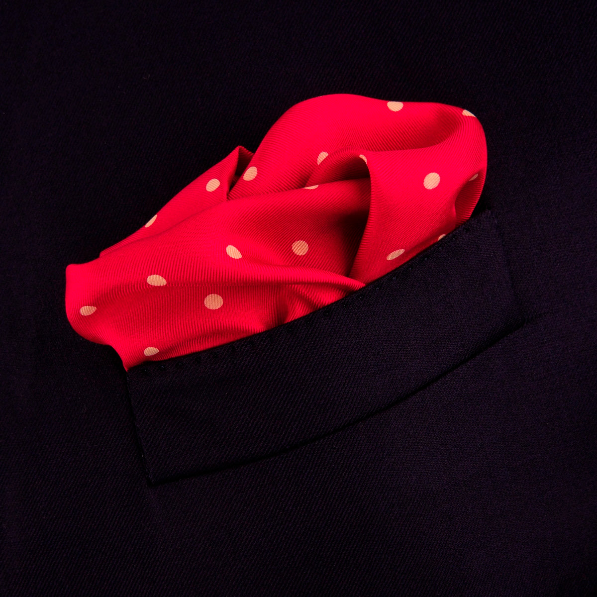 View 4 of product The Red and White Spot Silk Pocket Square by Turnbull & Asser features evenly spaced white polka dots on red silk with the brand's name printed in one corner. A folded edge reveals the reverse of this timeless pocket square.
