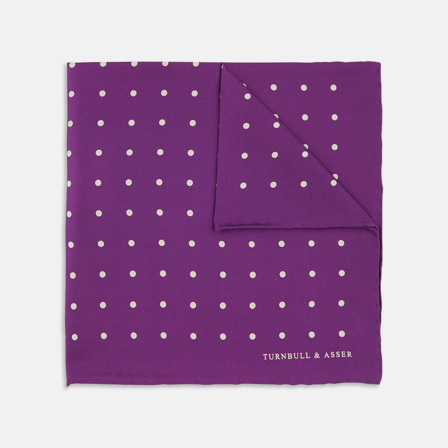 View 1 of product The Turnbull & Asser Purple Spot Silk Pocket Square features white polka dots and a small logo in the corner, with one corner folded over to showcase the underside.