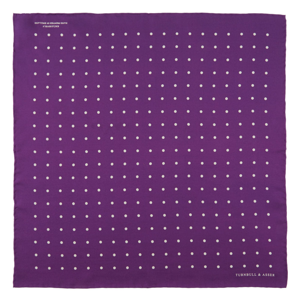 The Turnbull & Asser Purple Spot Silk Pocket Square features white polka dots and a small logo in the corner, with one corner folded over to showcase the underside.