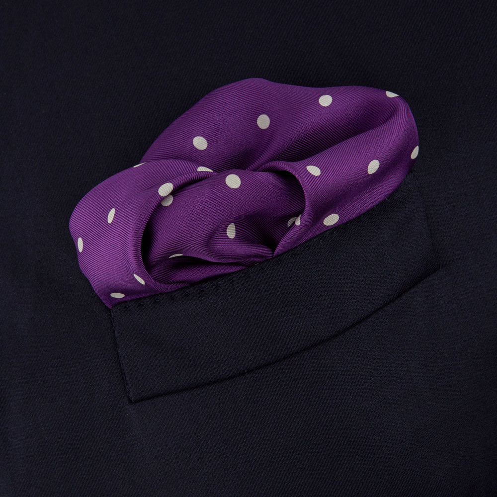 The Turnbull & Asser Purple Spot Silk Pocket Square features white polka dots and a small logo in the corner, with one corner folded over to showcase the underside.