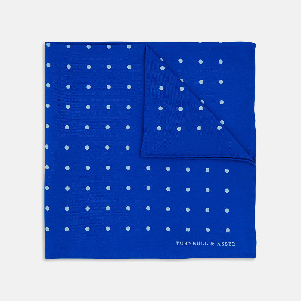 The Bright Blue Spot Silk Pocket Square by Turnbull & Asser features evenly spaced white polka dots and the brand's name printed in white in the bottom right corner, making it a stylish accessory for any suit.