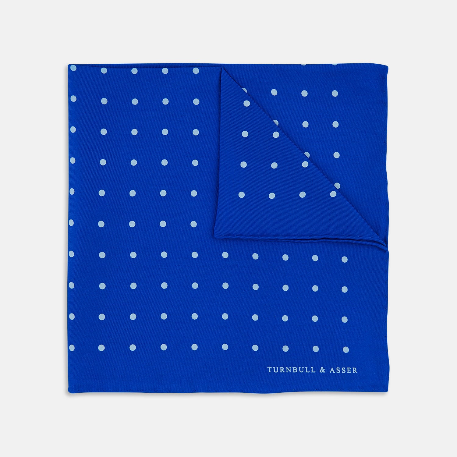 View 1 of product The Bright Blue Spot Silk Pocket Square by Turnbull & Asser features evenly spaced white polka dots and the brand's name printed in white in the bottom right corner, making it a stylish accessory for any suit.