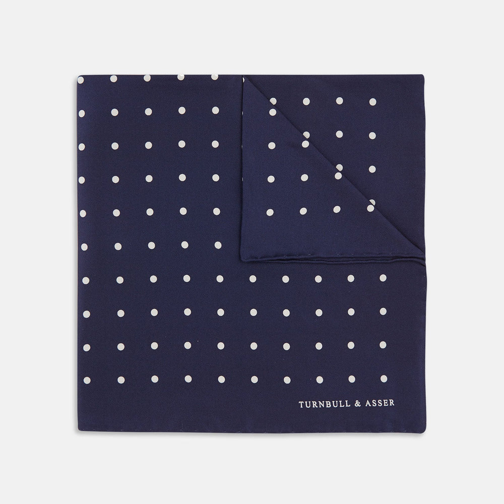 The Navy Spot Silk Pocket Square by Turnbull & Asser features a white polka dot pattern and is neatly folded at one corner, with the brand name printed in white on the lower right—perfect for pairing with pinstripe suits.