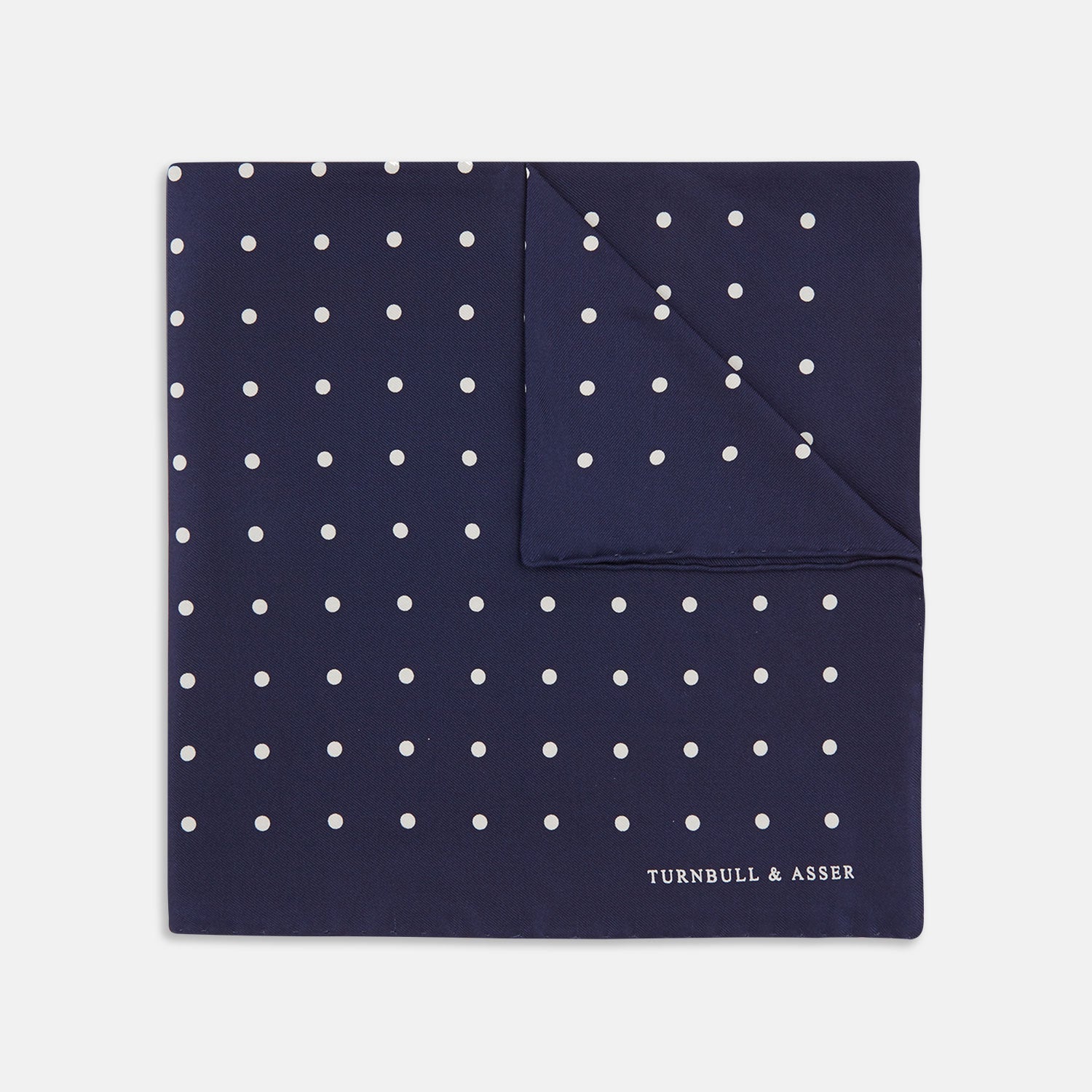 View 1 of product The Navy Spot Silk Pocket Square by Turnbull & Asser features a white polka dot pattern and is neatly folded at one corner, with the brand name printed in white on the lower right—perfect for pairing with pinstripe suits.