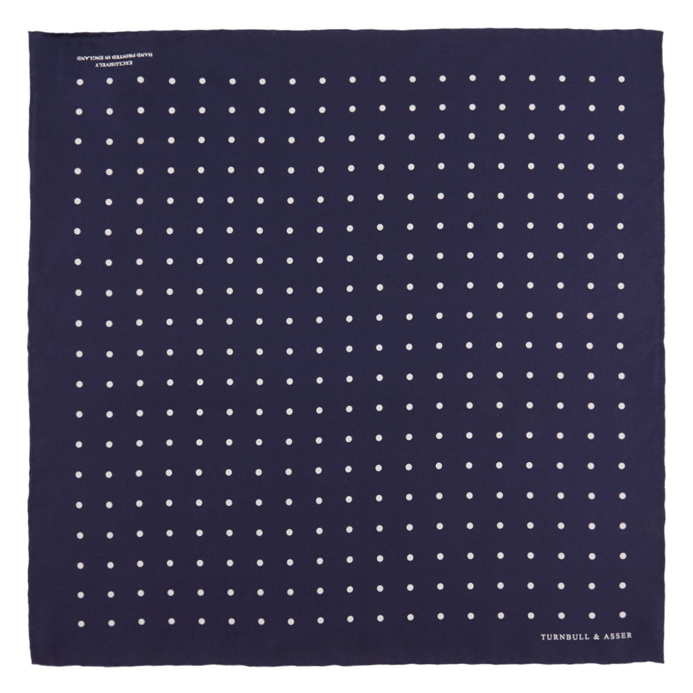 The Navy Spot Silk Pocket Square by Turnbull & Asser features a white polka dot pattern and is neatly folded at one corner, with the brand name printed in white on the lower right—perfect for pairing with pinstripe suits.