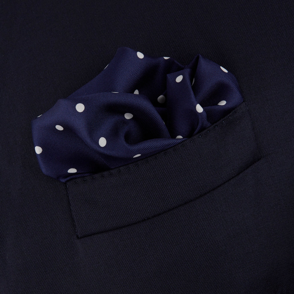 The Navy Spot Silk Pocket Square by Turnbull & Asser features a white polka dot pattern and is neatly folded at one corner, with the brand name printed in white on the lower right—perfect for pairing with pinstripe suits.