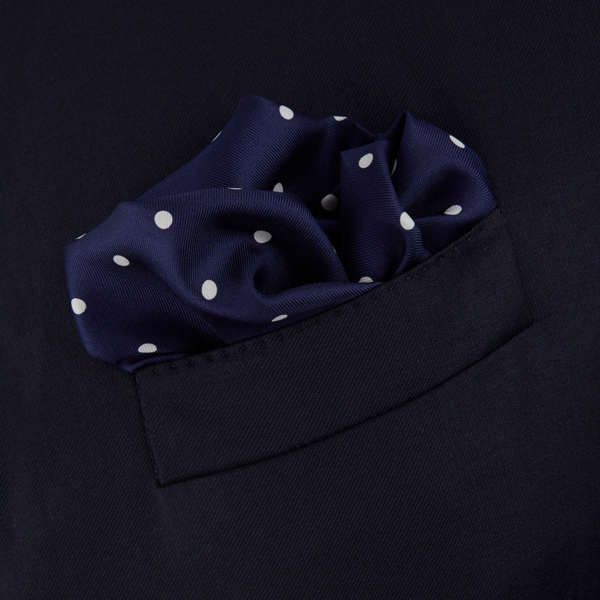 View 4 of product The Navy Spot Silk Pocket Square by Turnbull & Asser features a white polka dot pattern and is neatly folded at one corner, with the brand name printed in white on the lower right—perfect for pairing with pinstripe suits.