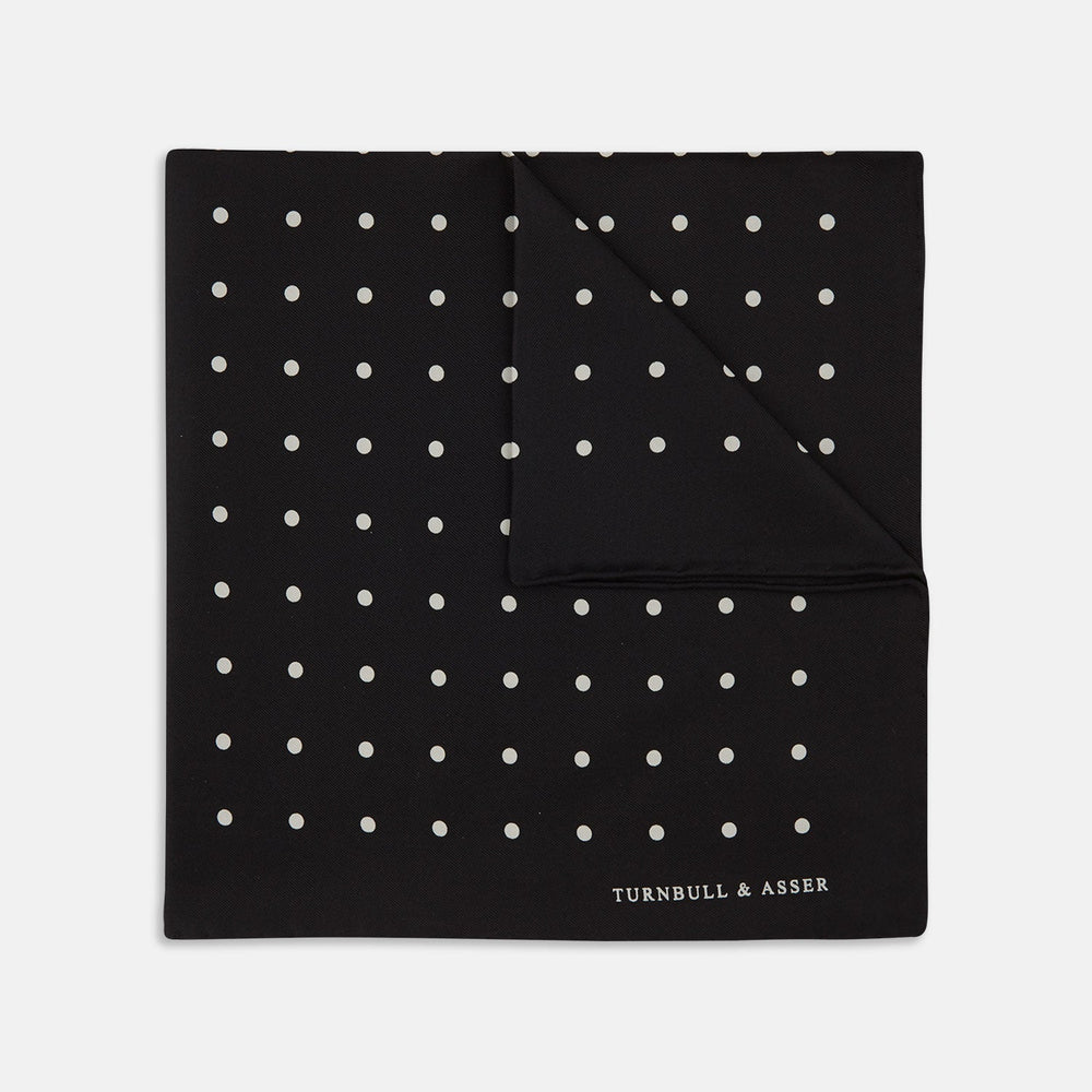 The Black Spot Silk Pocket Square by Turnbull & Asser features classic white polka dots on black silk, with the brand name printed in one corner and a stylish folded design.