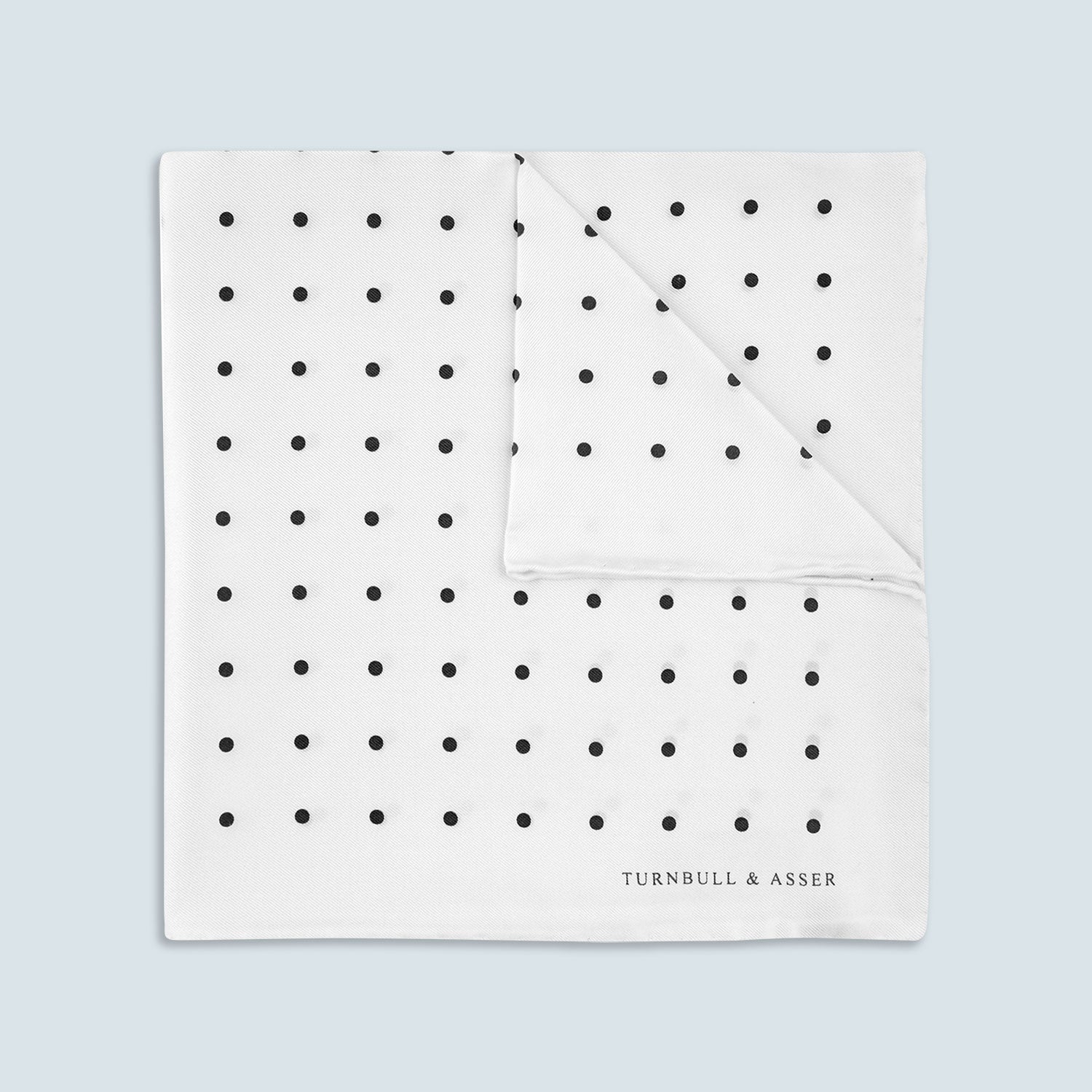 View 1 of product The Turnbull & Asser White and Black Spot Silk Pocket Square for men features evenly spaced black dots, a partial corner fold, and the brand name printed in black in the lower right corner.