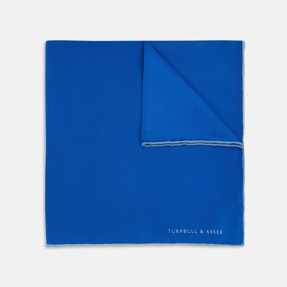 The Royal Blue Piped Silk Pocket Square by Turnbull & Asser features a vibrant blue hue, subtle stitched border, and the brand name printed in white in the lower right corner.