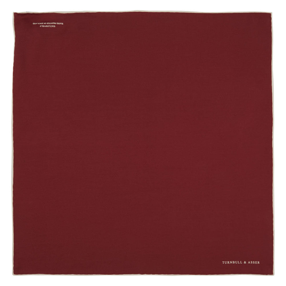 The Turnbull & Asser Burgundy and White Piped Silk Pocket Square features a classic burgundy design with a subtle white piped edge and the brand's logo printed in white on the bottom right corner.