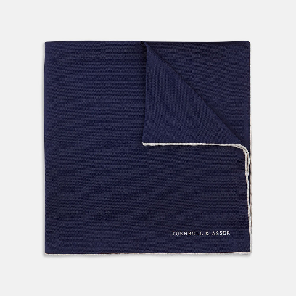 The Navy and White Piped Silk Pocket Square by Turnbull & Asser features white edging and partial corner folding, with the brand’s name printed in white near the lower edge.