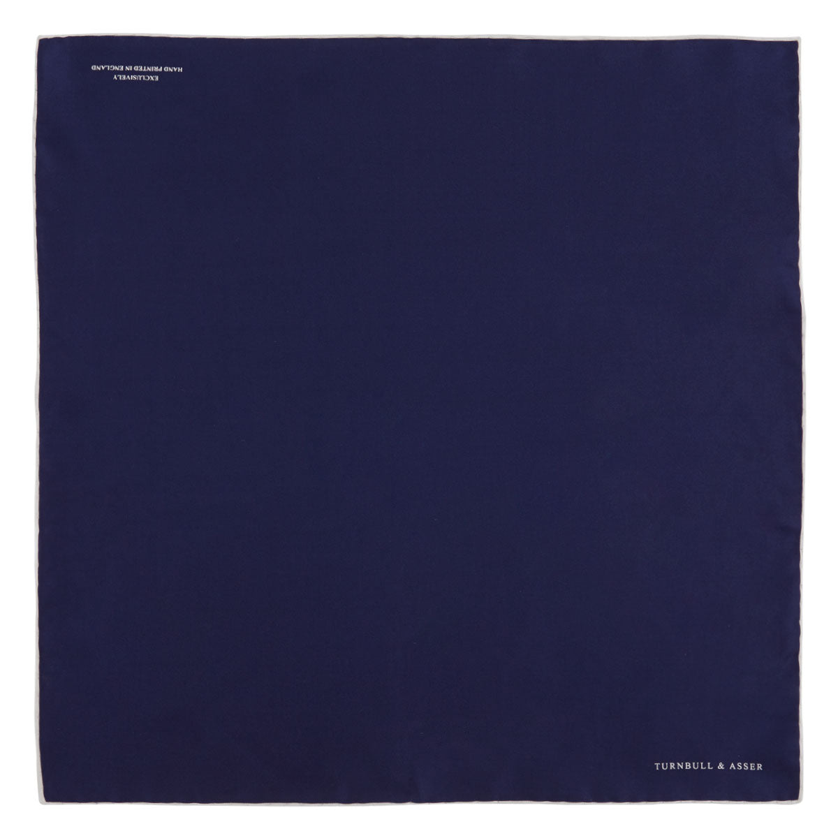 View 2 of product The Navy and White Piped Silk Pocket Square by Turnbull & Asser features white edging and partial corner folding, with the brand’s name printed in white near the lower edge.