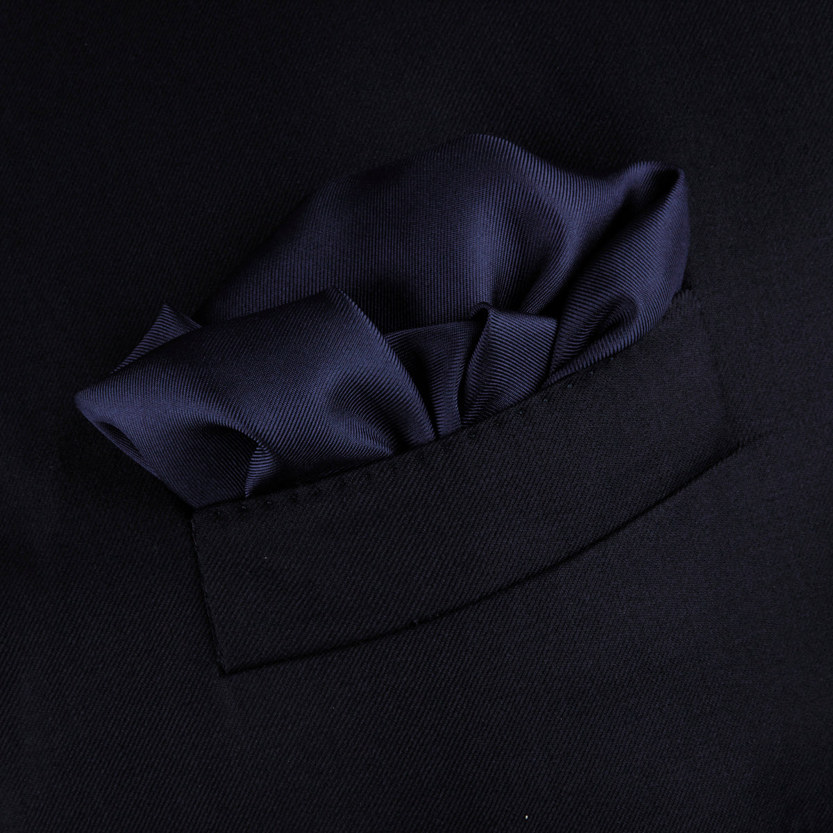 View 4 of product The Navy and White Piped Silk Pocket Square by Turnbull & Asser features white edging and partial corner folding, with the brand’s name printed in white near the lower edge.