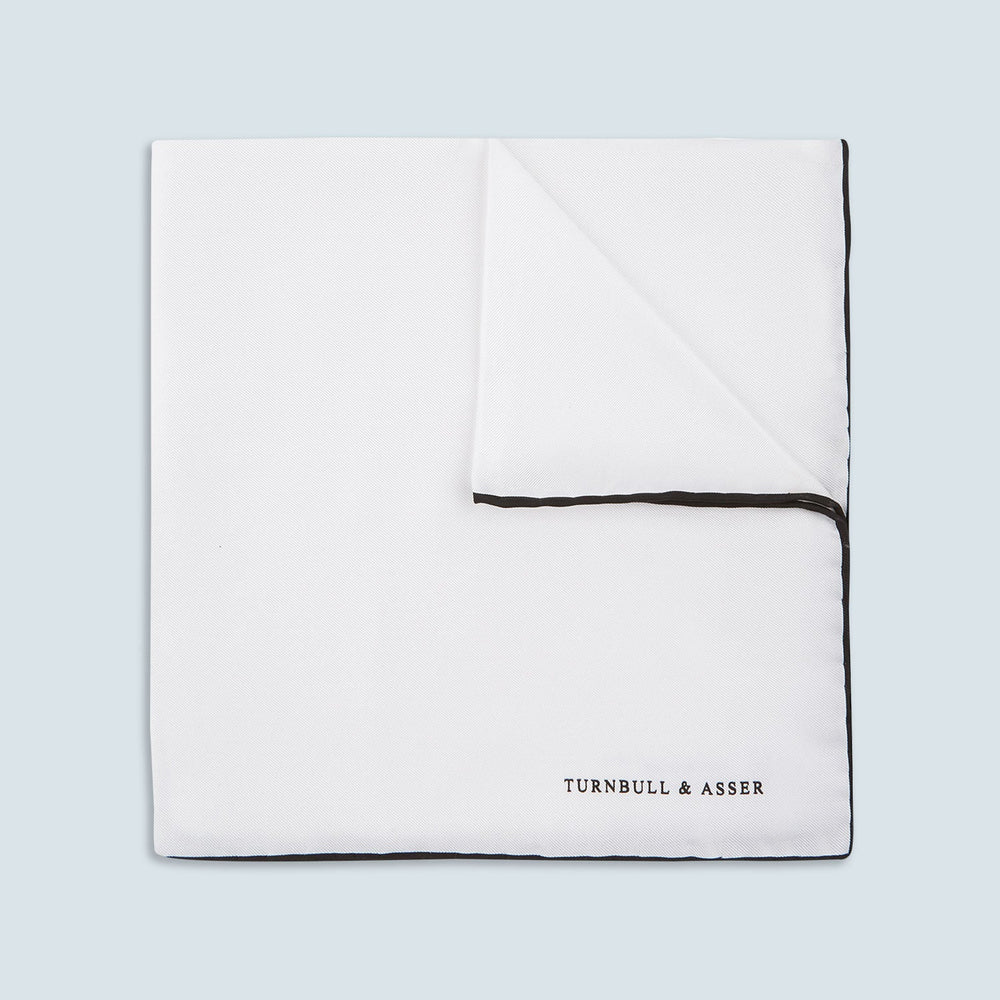 The Turnbull & Asser White and Black Piped Silk Pocket Square features a crisp white base with black edging, neatly folded to showcase the logo in black on the lower right—elegant monochrome style for any occasion, shown on a light gray background.