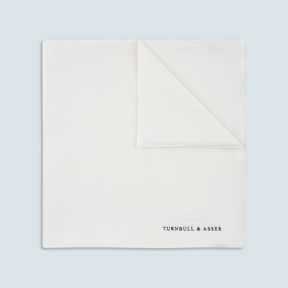 The White Piped Silk Pocket Square by Turnbull & Asser is shown folded at one corner with the brand's logo in black on the lower right, set against a light blue background.