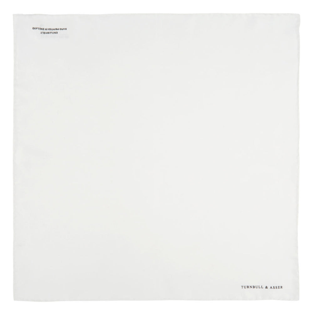 The White Piped Silk Pocket Square by Turnbull & Asser is shown folded at one corner with the brand's logo in black on the lower right, set against a light blue background.
