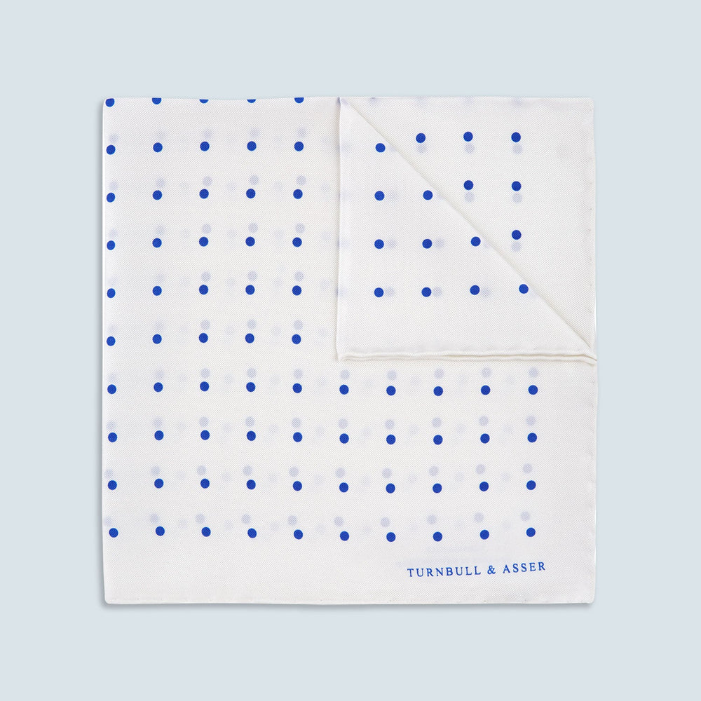 The Turnbull & Asser White and Blue Spot Silk Pocket Square features evenly spaced blue dots, a brand logo in one corner, and is displayed on a light blue background with a stylishly folded corner.