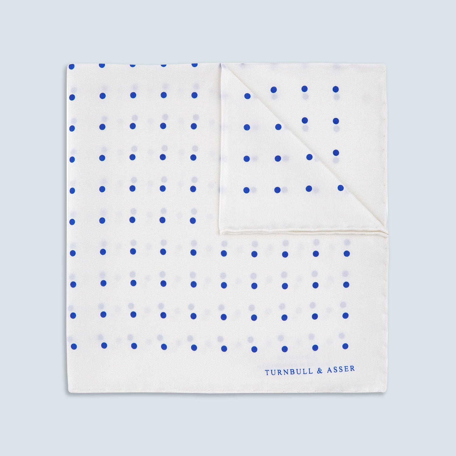 View 1 of product The Turnbull & Asser White and Blue Spot Silk Pocket Square features evenly spaced blue dots, a brand logo in one corner, and is displayed on a light blue background with a stylishly folded corner.