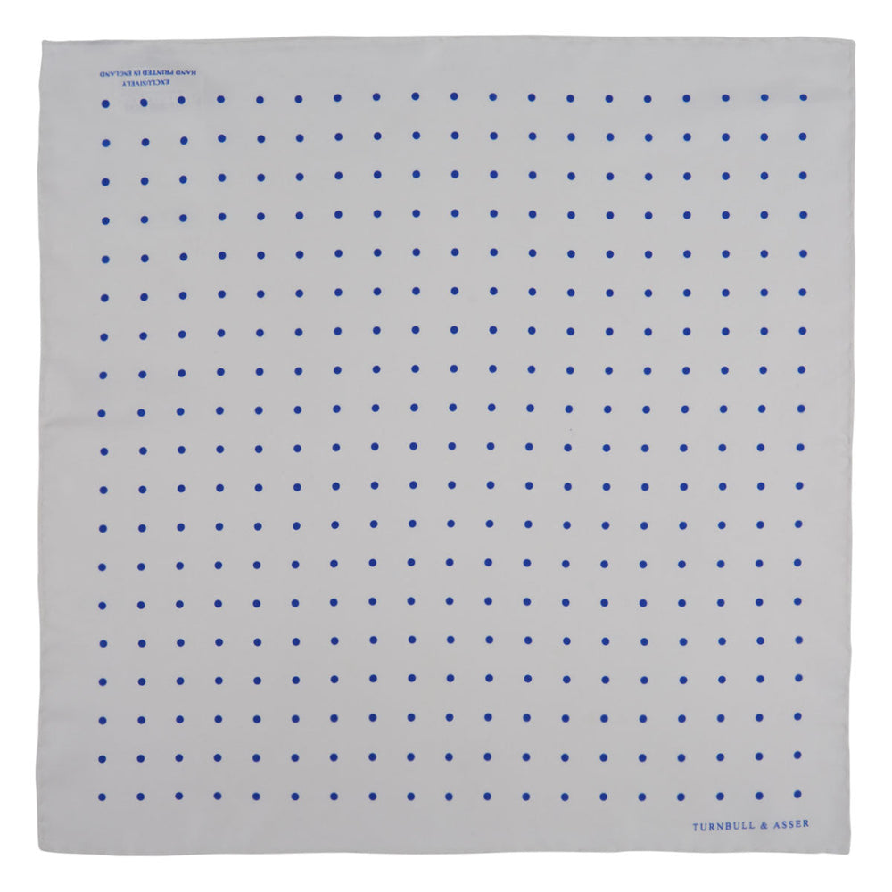 The Turnbull & Asser White and Blue Spot Silk Pocket Square features evenly spaced blue dots, a brand logo in one corner, and is displayed on a light blue background with a stylishly folded corner.