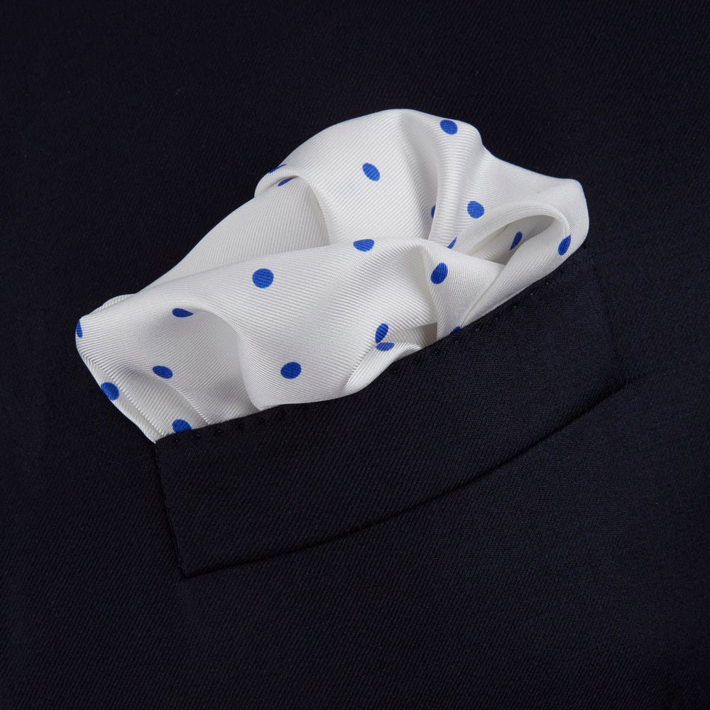The Turnbull & Asser White and Blue Spot Silk Pocket Square features evenly spaced blue dots, a brand logo in one corner, and is displayed on a light blue background with a stylishly folded corner.
