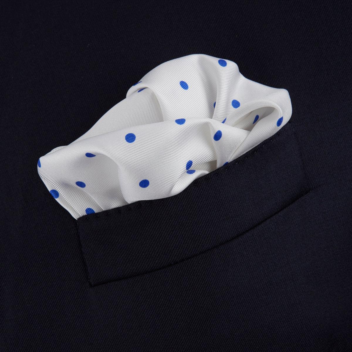 View 4 of product The Turnbull & Asser White and Blue Spot Silk Pocket Square features evenly spaced blue dots, a brand logo in one corner, and is displayed on a light blue background with a stylishly folded corner.