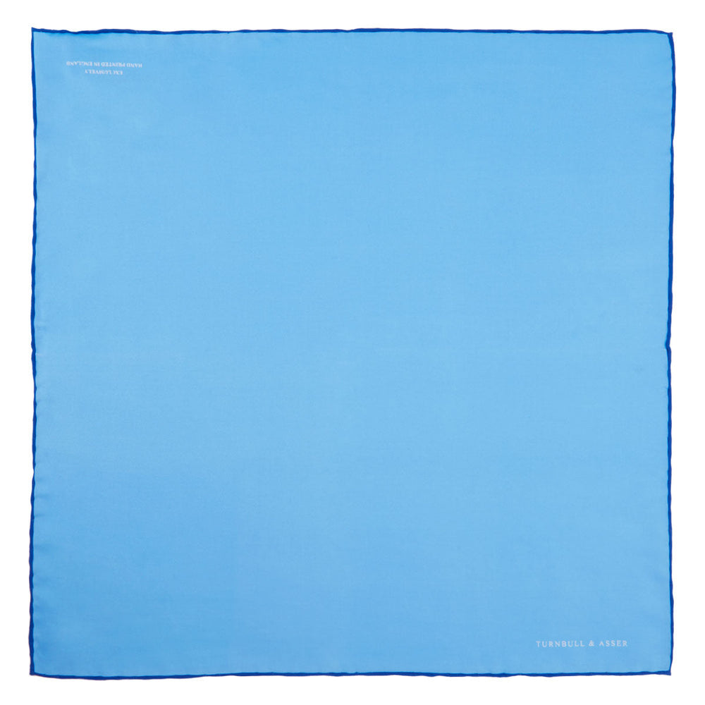 The Light Blue and Navy Piped Silk Pocket Square by Turnbull & Asser, neatly folded with dark blue edges and the brand’s white logo in the corner, is shown on a plain white background—a refined accessory for any occasion.