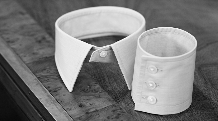 A black-and-white photo shows a detached white dress shirt collar upright next to a matching detached shirt cuff with three buttons, both resting on a wooden surface.