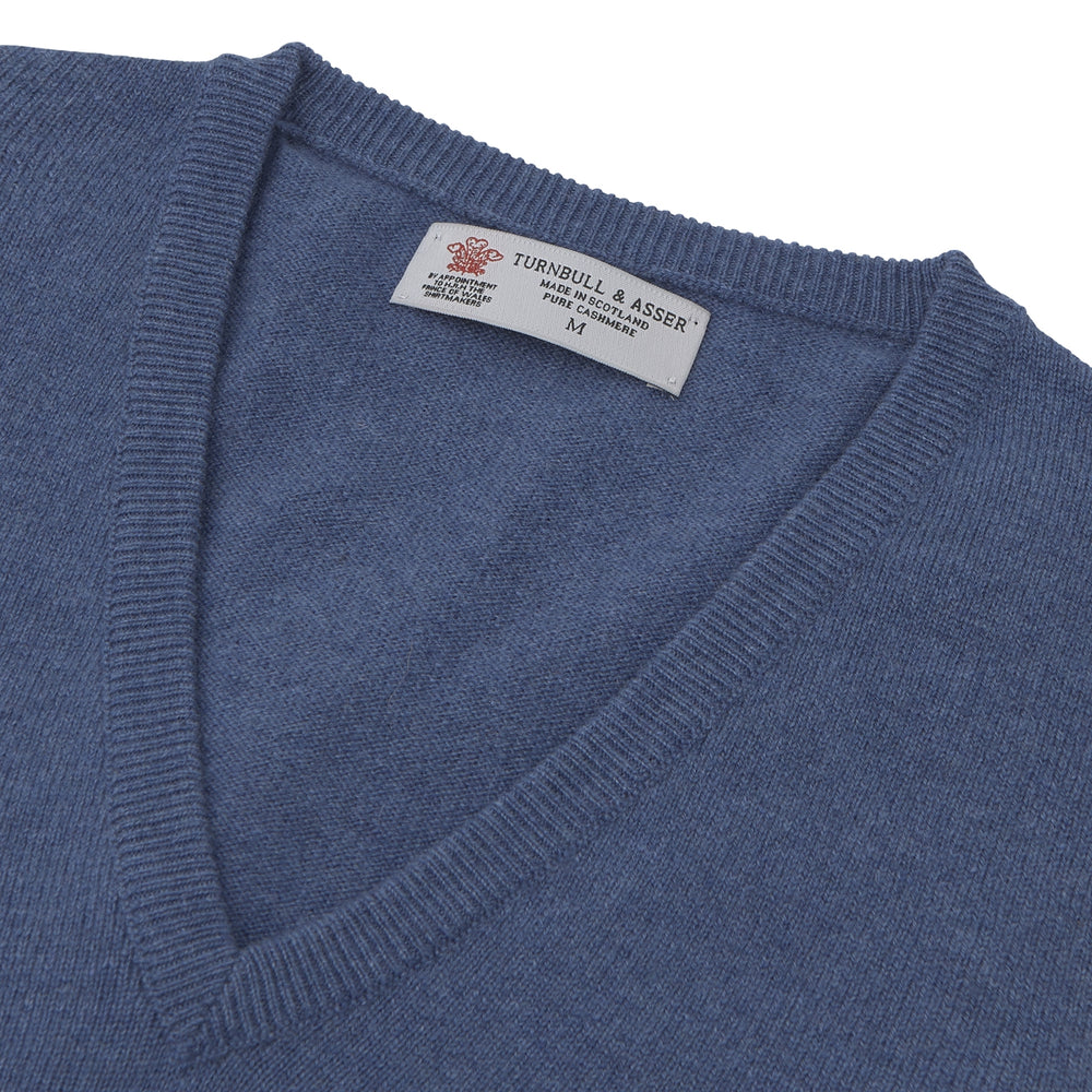 The Turnbull & Asser Blue V-Neck Cashmere Slipover, neatly folded with the label visible, is crafted from soft cashmere and ideal for formal layering. Shown against a white background.