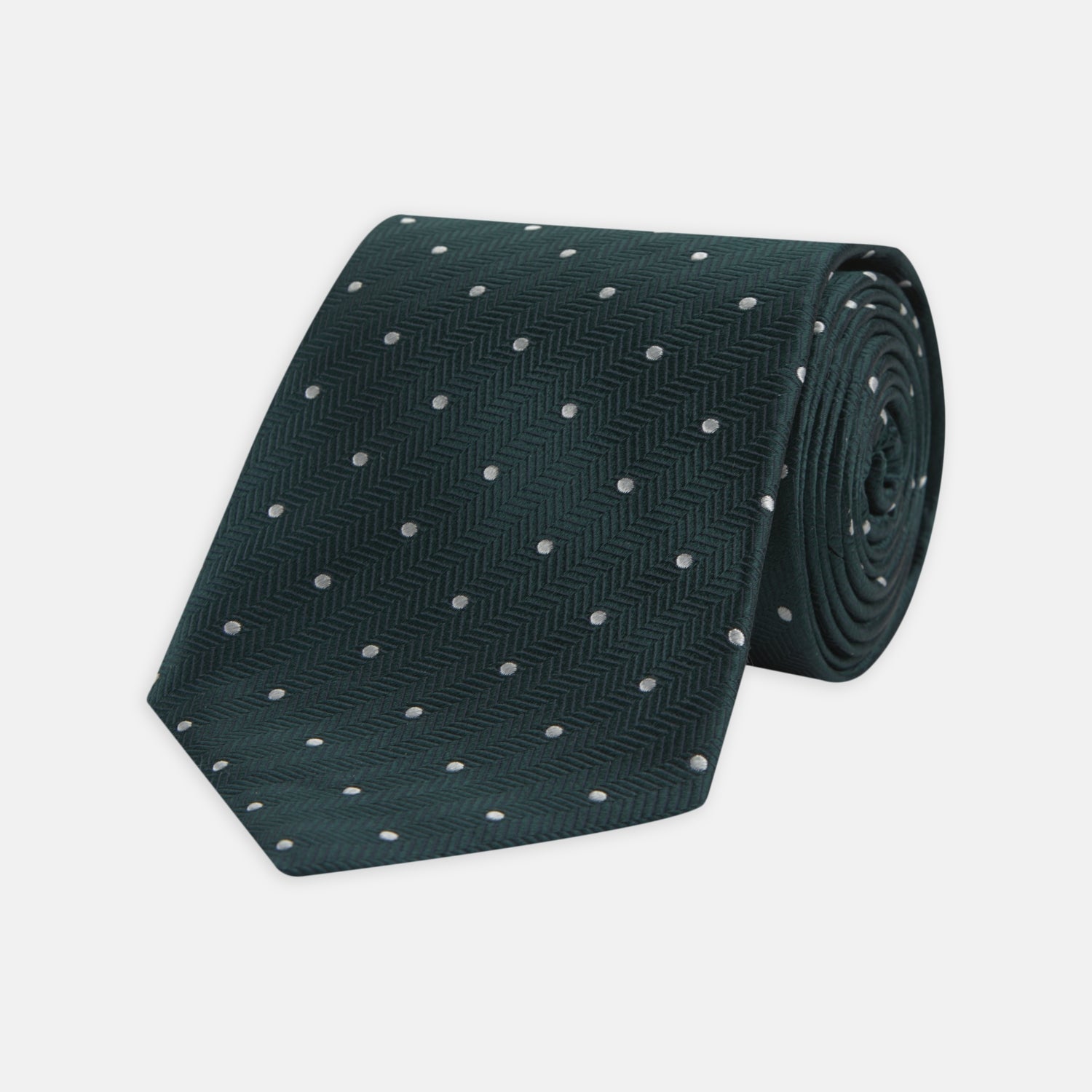 View 1 of product The Forest Green and White Small Spot Herringbone Silk Tie by Turnbull & Asser features a dark green woven herringbone pattern with small white spots, crafted from fine English silk and shown rolled up on a white background.