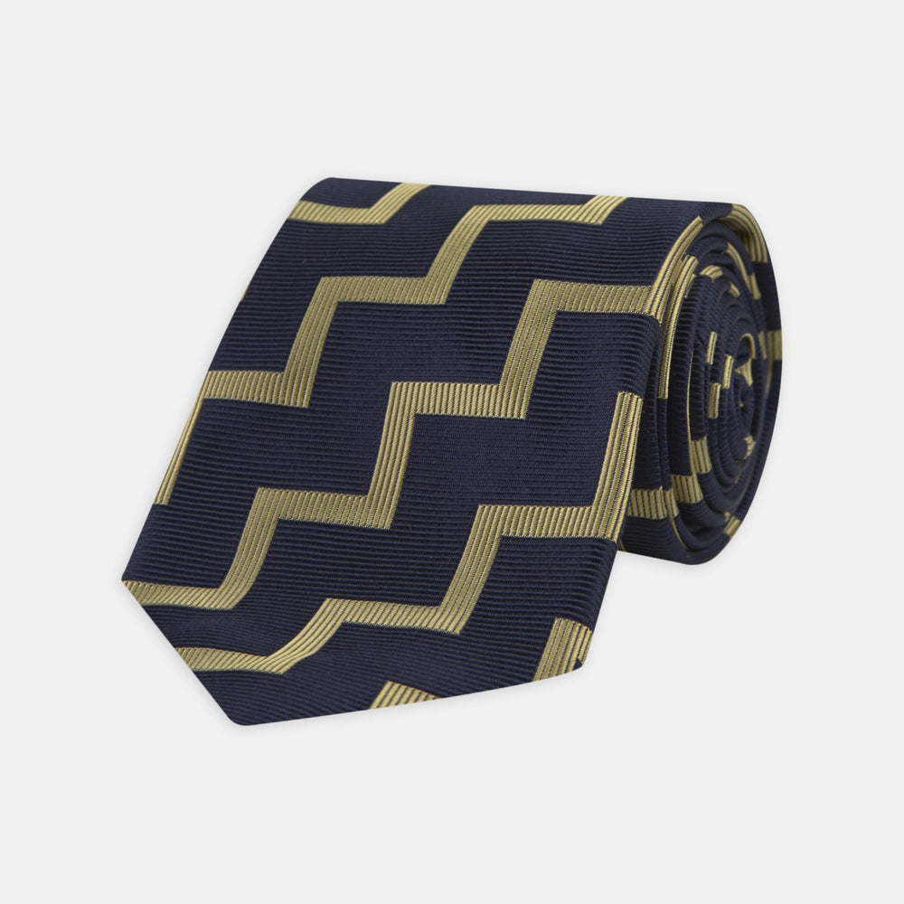 The Navy and Gold Striped Zigzag Silk Tie by Turnbull & Asser features a bold geometric pattern—an essential modern accessory shown rolled up with the pointed end forward on a white background.