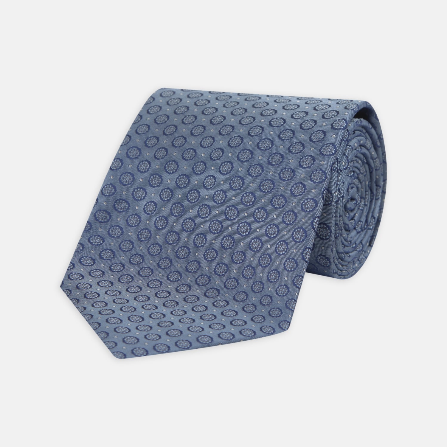 View 1 of product The Light Blue Floral Spot Silk Tie by Turnbull & Asser features a classic pattern in varied blue tones, rolled up on a white background—an ideal accessory to complement any office shirt.