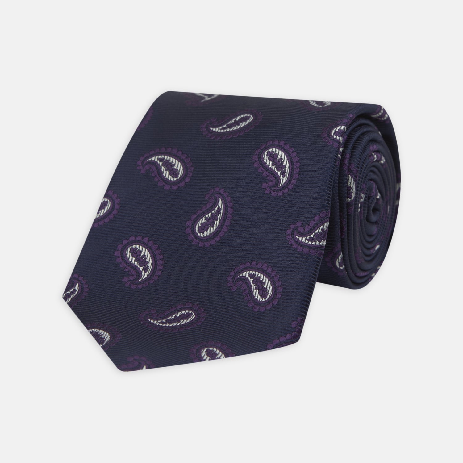 View 1 of product The Turnbull & Asser Navy and Plum Floating Paisley Silk Tie features a repeating purple and white paisley pattern, elegantly rolled up on a white background.