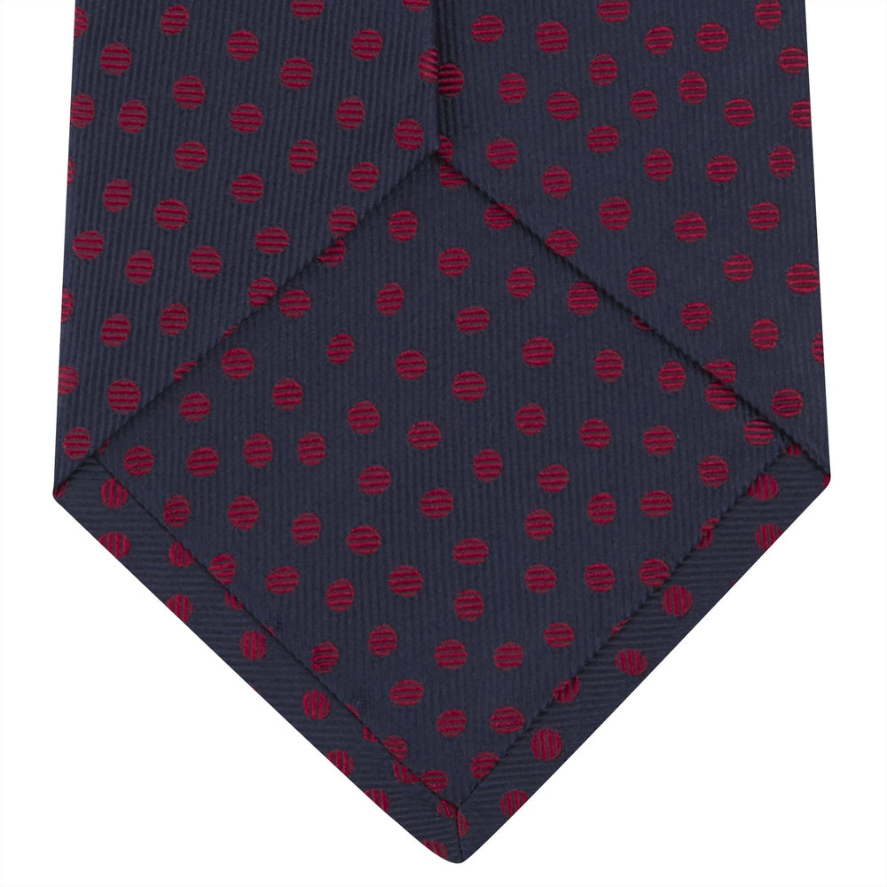 The Turnbull & Asser Navy and Red Paint Spot Silk Tie, featuring red spots on navy blue silk, is rolled with the wide end forward—ideal for office shirts and shown against a white background.