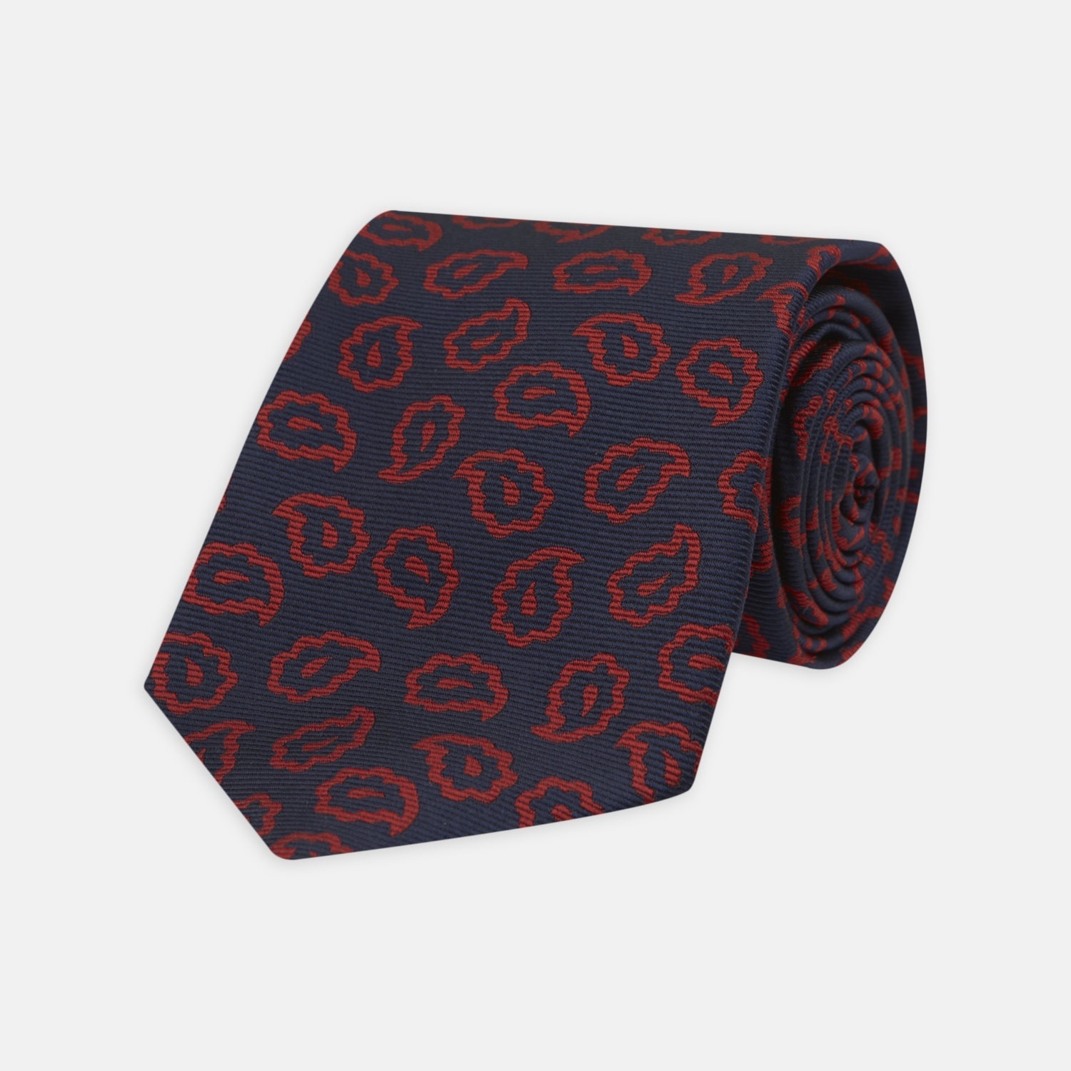 View 1 of product The Navy and Red Petal Paisley Silk Tie by Turnbull & Asser features an elegant paisley pattern on navy silk, rolled at one end and displayed on a plain white background.