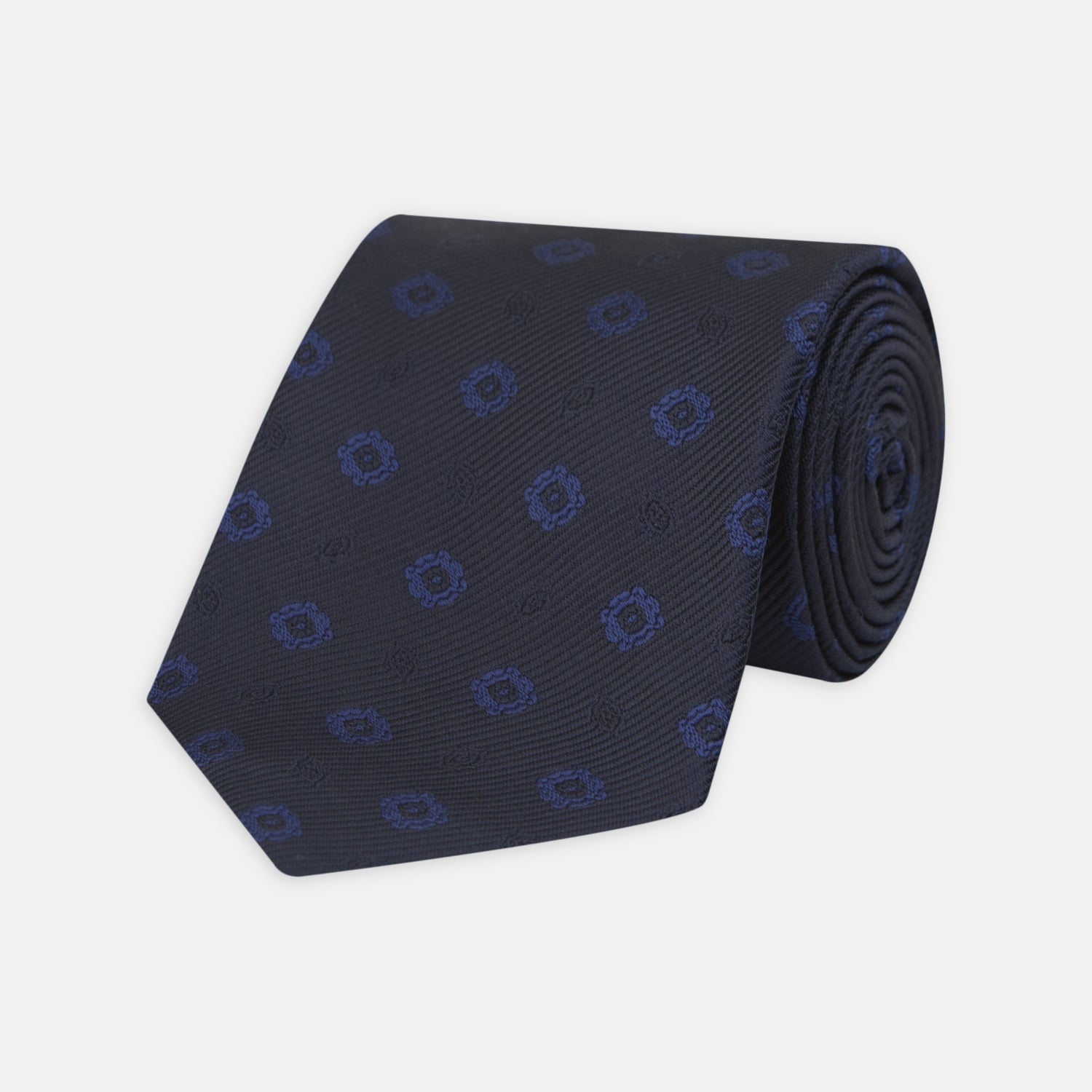 View 1 of product The Turnbull & Asser Navy and Blue Emblem Spot Silk Tie features a classic pattern of light blue motifs on navy silk, showcased rolled on a white background—a refined accessory for any office shirt.